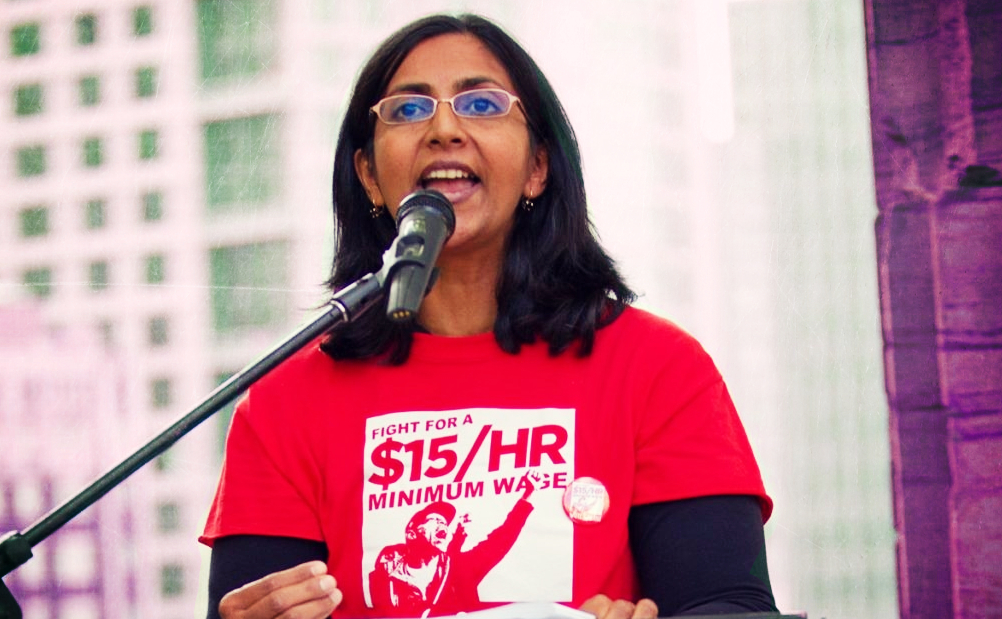 Photo of Kashama Sawant speaking from a podium, an urban tower is the background.