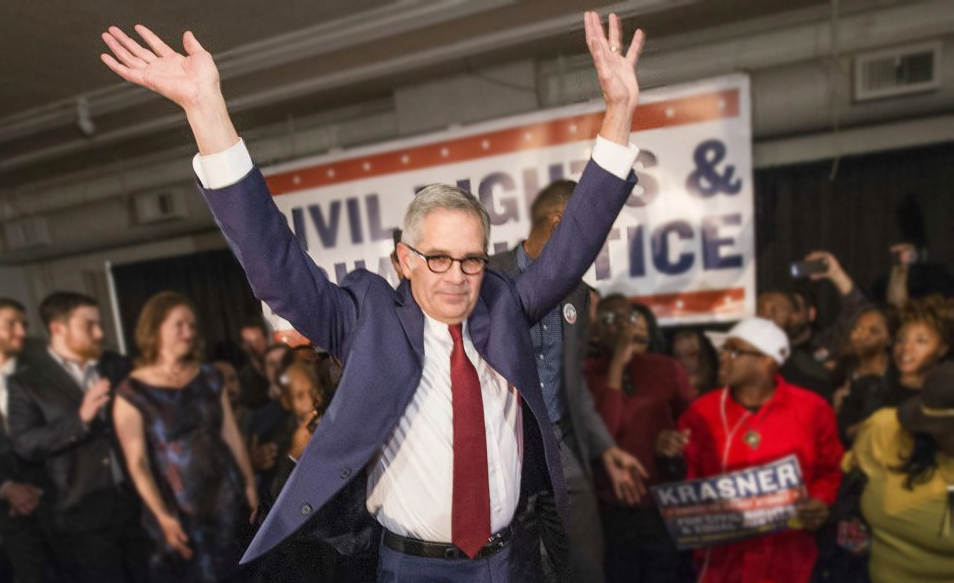 Philadelphia District Attorney Larry Krasner celebrates at a campaign event, hands in air and supporters behind him. 