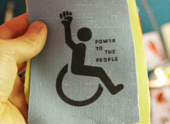 Photo of a screen printed patch with outline of wheel chair symbol with raised fist and words "Power to the people."