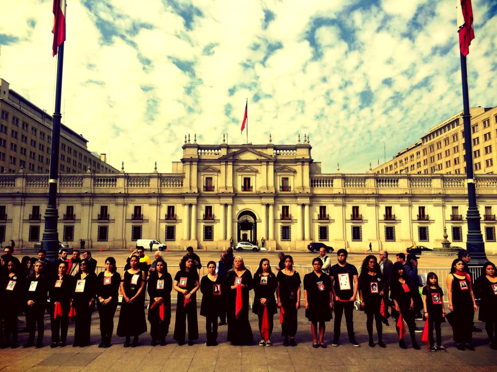 Groups of individuals dressed in black standing in line with government building in background. Pictured is the “Cueca Sola” Collective outside of the Casa Moneda, the national palace in Santiago, Chile, which performs the Chilean national dance, known as "cueca," which is always done with a partner. In remembrance the performances is done alone, "sola," for one or more of the disappeared, often a friend or family member. Taken September 11, 2018. 