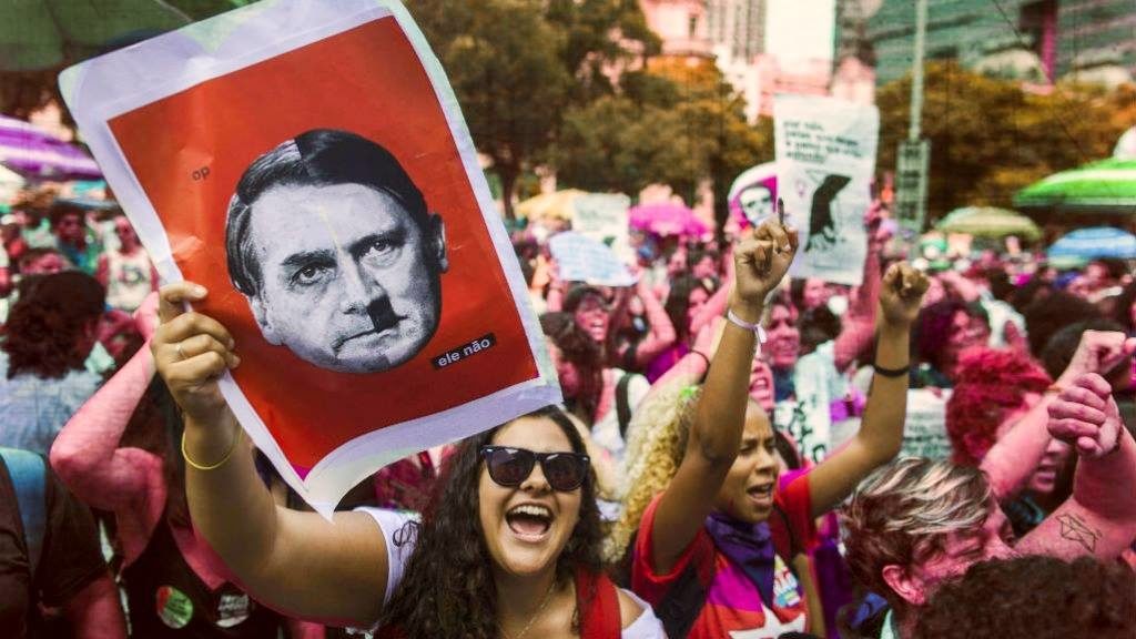 Crowd of protesters, woman holding sign depicting Bolsonaro in the likeness of Hitler