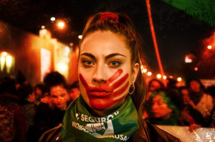 Image of woman at protest during evening. A hand painted in red is on her face, symbolic of deaths resulting from illegal abortion.