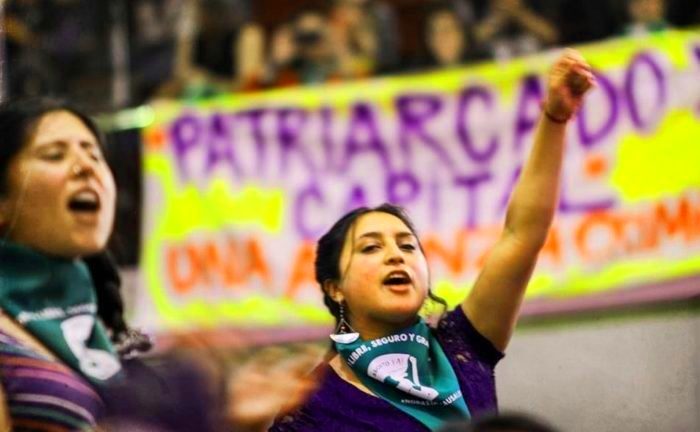 Left side: woman chanting, center: woman with fist in air. Both are wearing green bandanas which are the symbol of the feminist abortion rights movements. Background: banner denouncing capitalist patriarchy. 