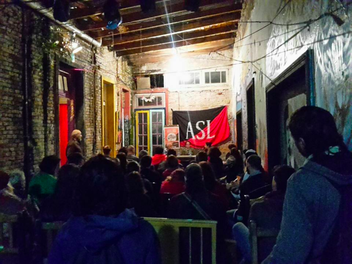 Seated crowd facing towards a presentation, wall has large red and black flag with "ASL."