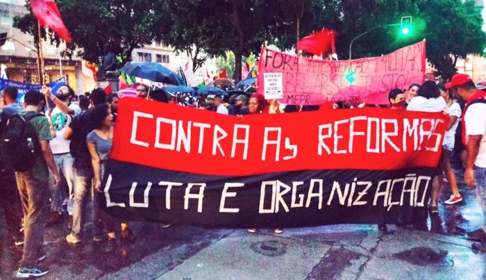 Red and black banner being held during a demonstration reads in Portuguese "Against the reforms / Struggle and organize"