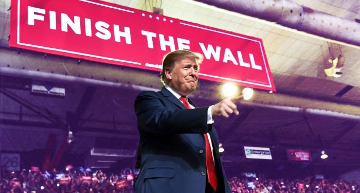 Trump on stage at a campaign rally pointing towards audience, large banner in background "finish the wall."
