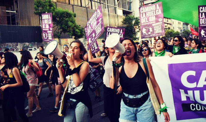 Women's march in Chile. Two women in foreground lead chants with bull horns. Signs say "camino a la huelga del 8m / on the road to strike March 8"