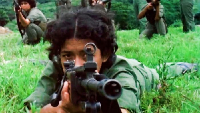 Screen shot from Las Sandinistas film showing woman guerrilla laying in grass with military rifle pointing towards viewer.  